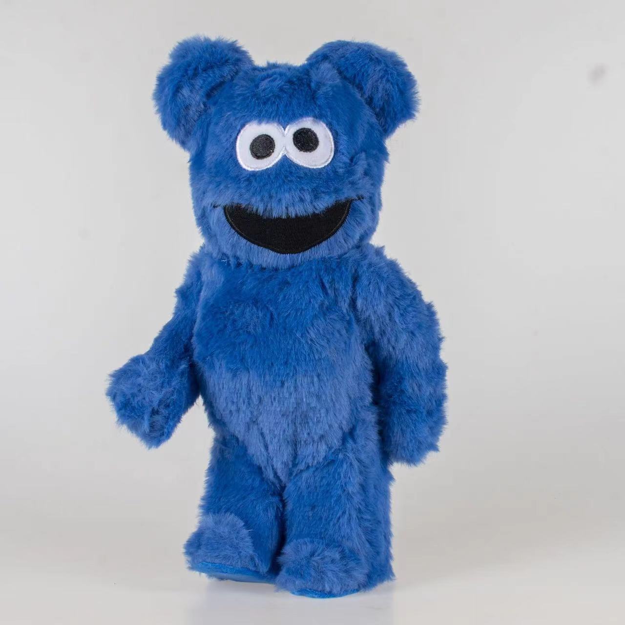 A blue furry character plush toy standing against a plain light gray background. The plush has large white eyes with black pupils, a wide black smile, and short rounded ears.