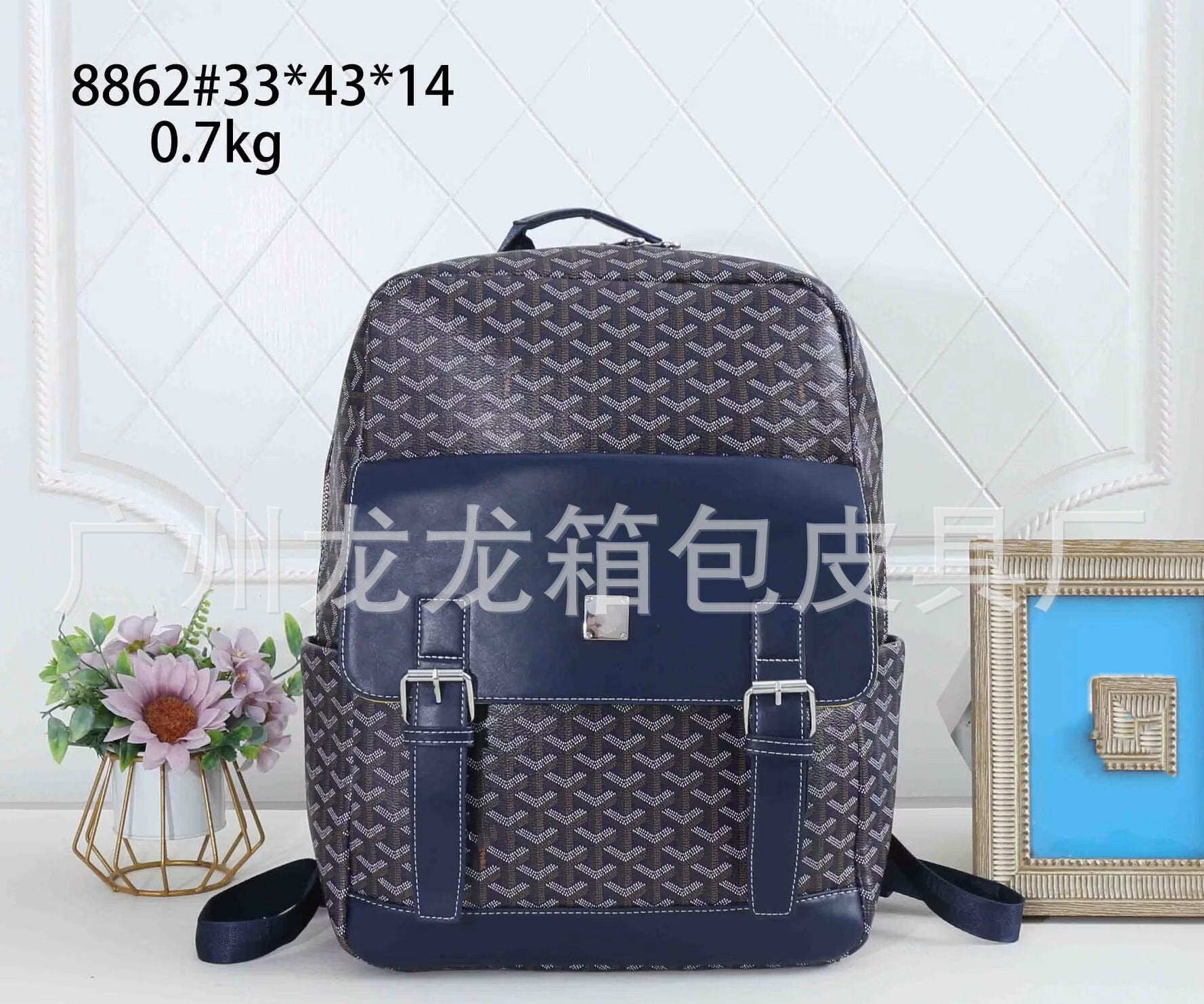A dark gray and navy blue patterned backpack with a navy blue front flap and buckle straps, displayed on a white surface with decorative props including a small flower vase and a picture frame. Text overlays show product details and a shop URL.