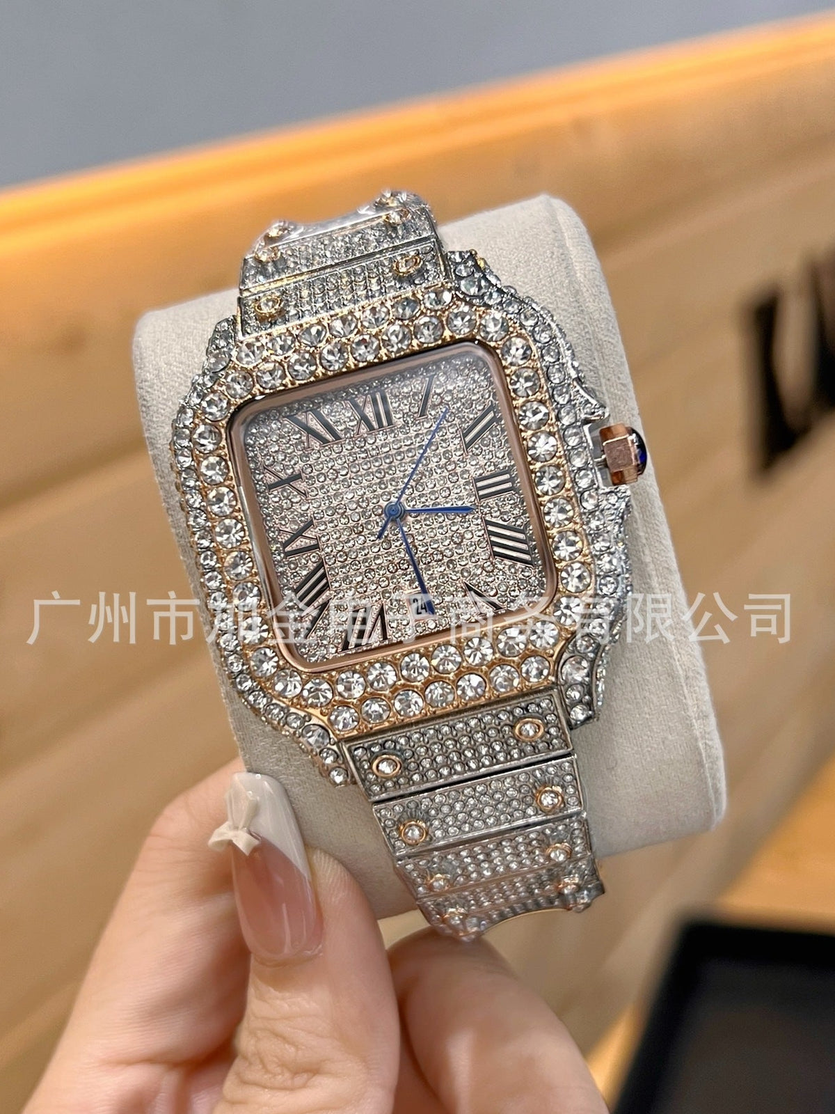 A heavily embellished rectangular watch with a diamond-covered silver-toned case and matching crystal-encrusted bracelet, held in a hand against a blurred wooden background.