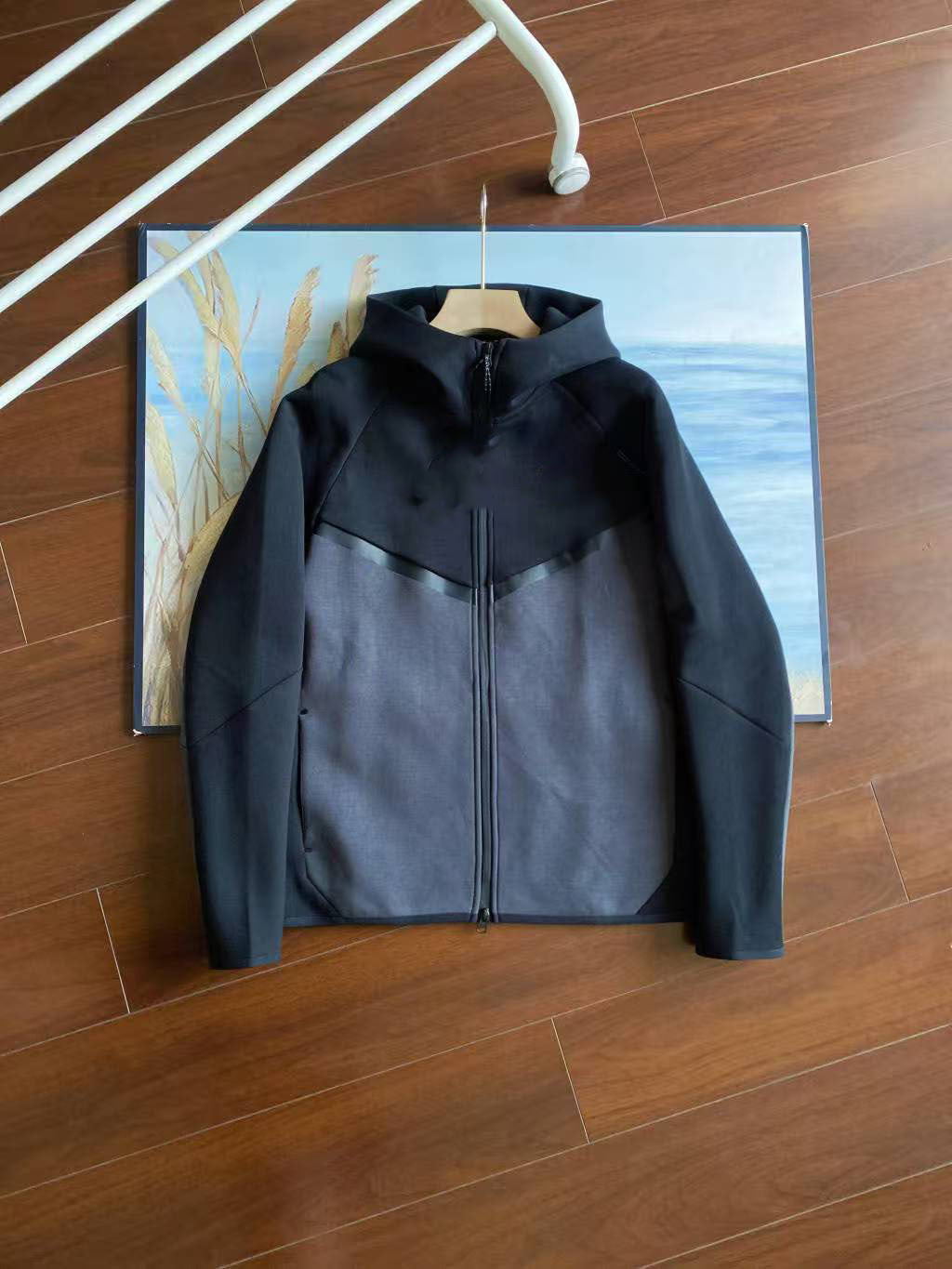 A colorblock zip-up hooded jacket in black and dark gray, hanging on a wooden hanger against a framed art print on a wooden floor.