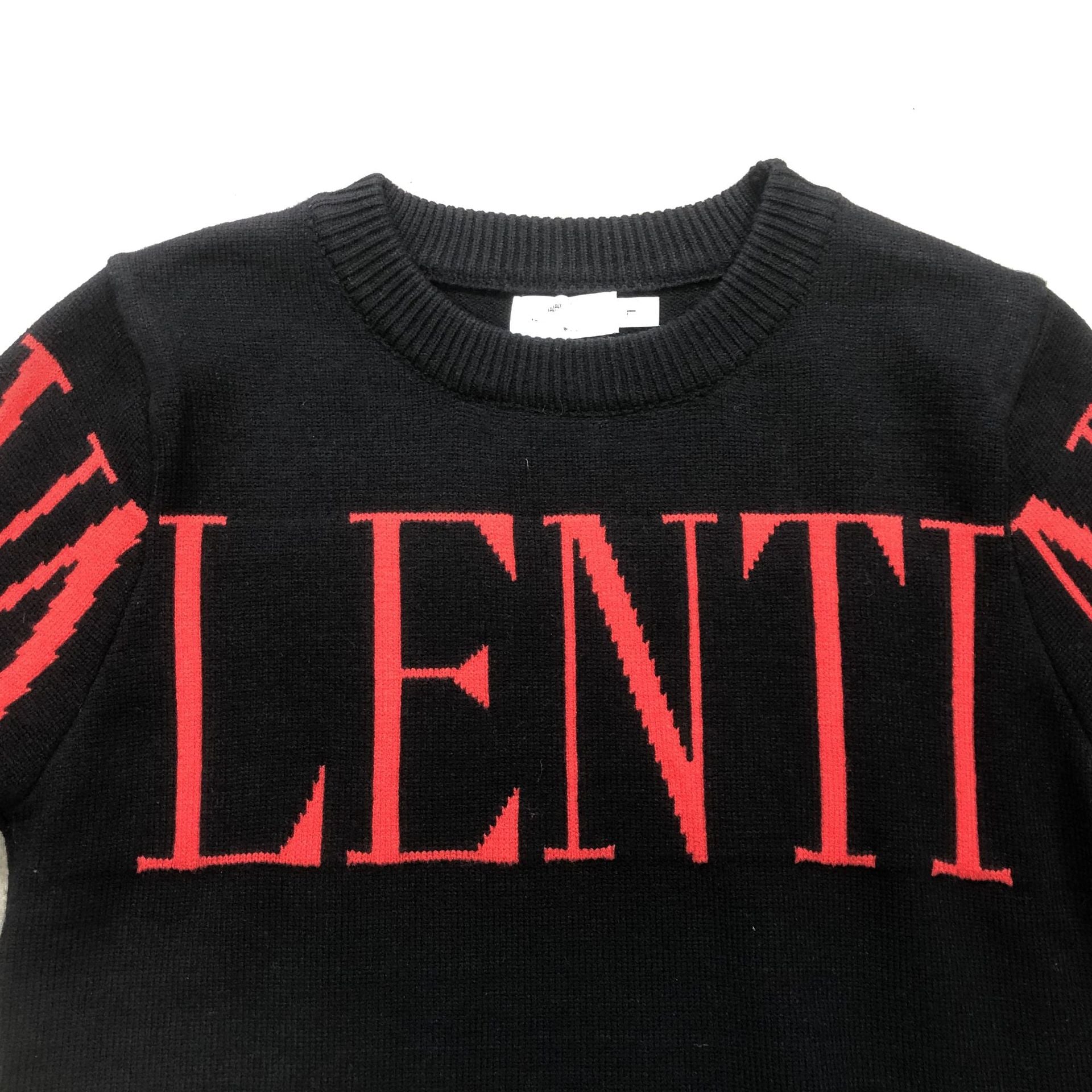 A close-up of a black knit sweater featuring the word 'VALENTI' in large red letters across the chest, with red graphic details on the sleeves, set against a plain white background.