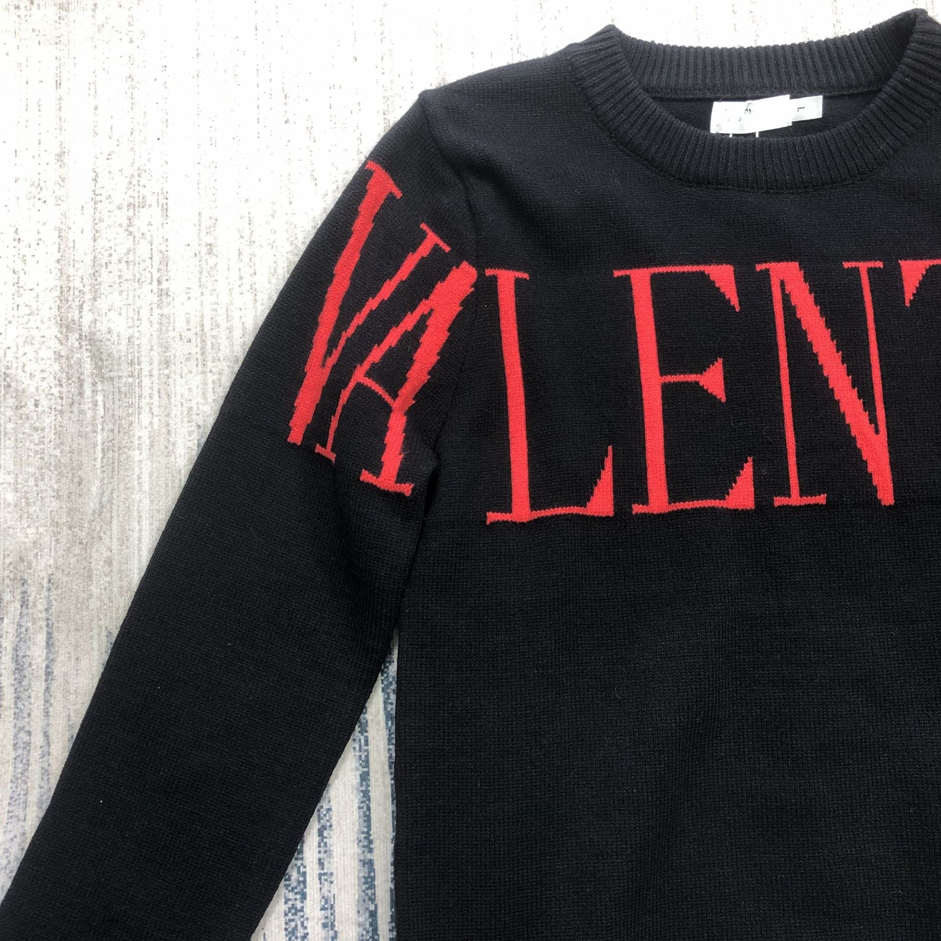 A close-up of a black knit sweater with a bold red 'VALENTI' lettering across the chest, laid flat on a textured light gray and white surface.