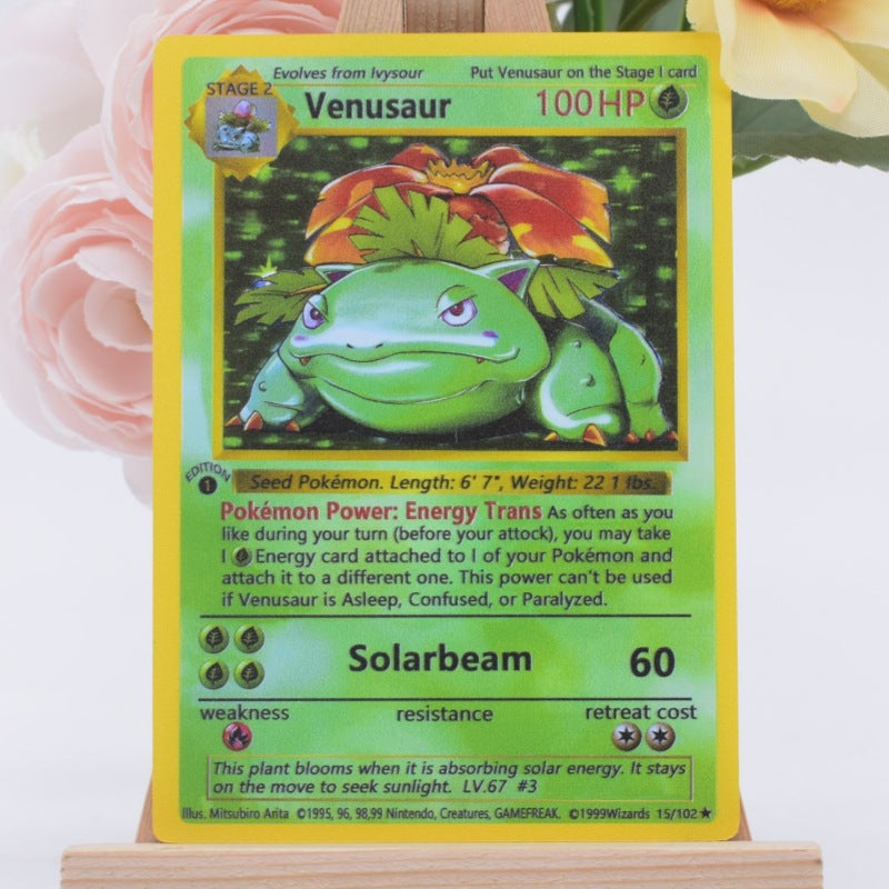 A Venusaur Pokémon trading card displayed upright on a small wooden stand, with soft pink and yellow flowers blurred in the background.