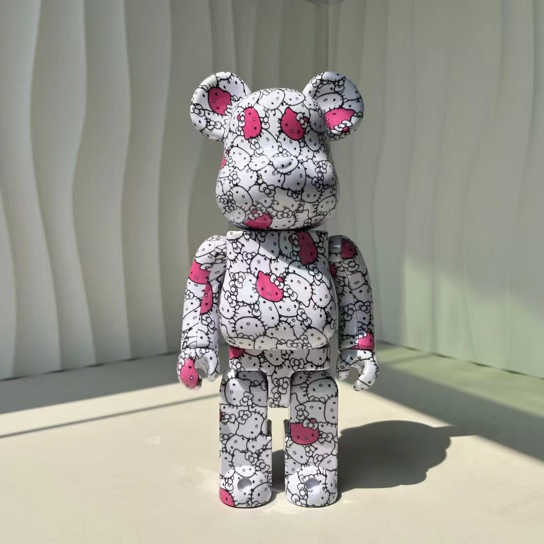 A bear-shaped collectible figure standing upright, covered in a white and black repeating Hello Kitty-style face pattern with bright pink bow accents, photographed against a pale draped backdrop.