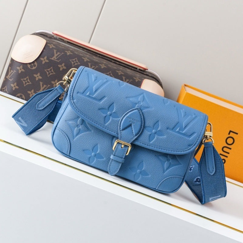 A blue Louis Vuitton monogram embossed leather shoulder bag with gold-tone hardware, displayed on a white shelf next to a Louis Vuitton box and a brown monogram pouch.