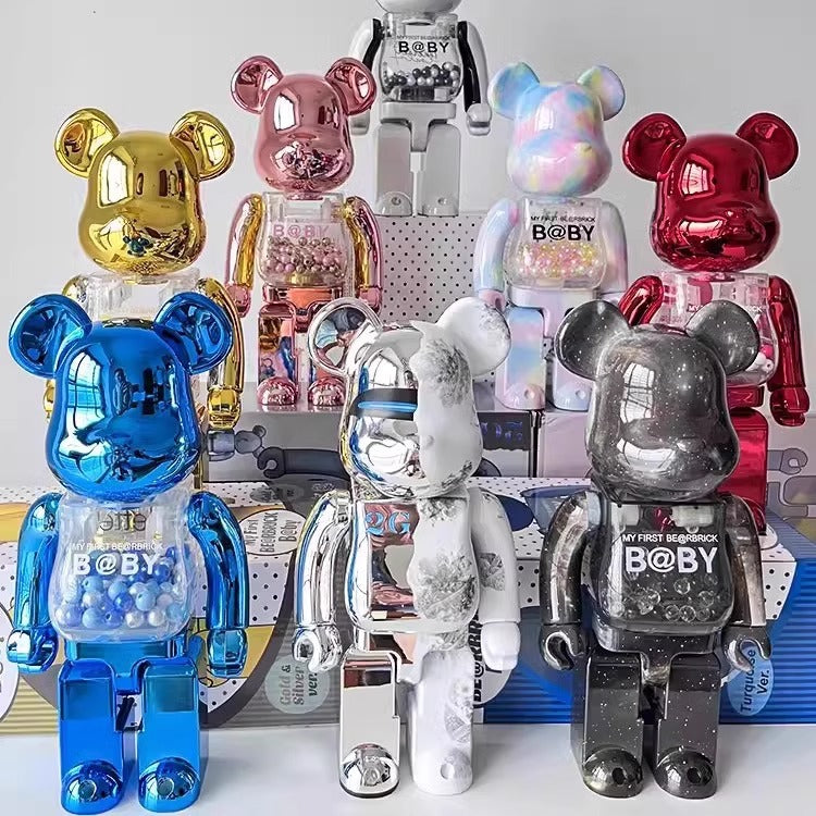 A group of BE@BER Bearbrick figures in various metallic and glitter finishes, including gold, pink, iridescent, red, blue, white, and dark gray, arranged on display stands.