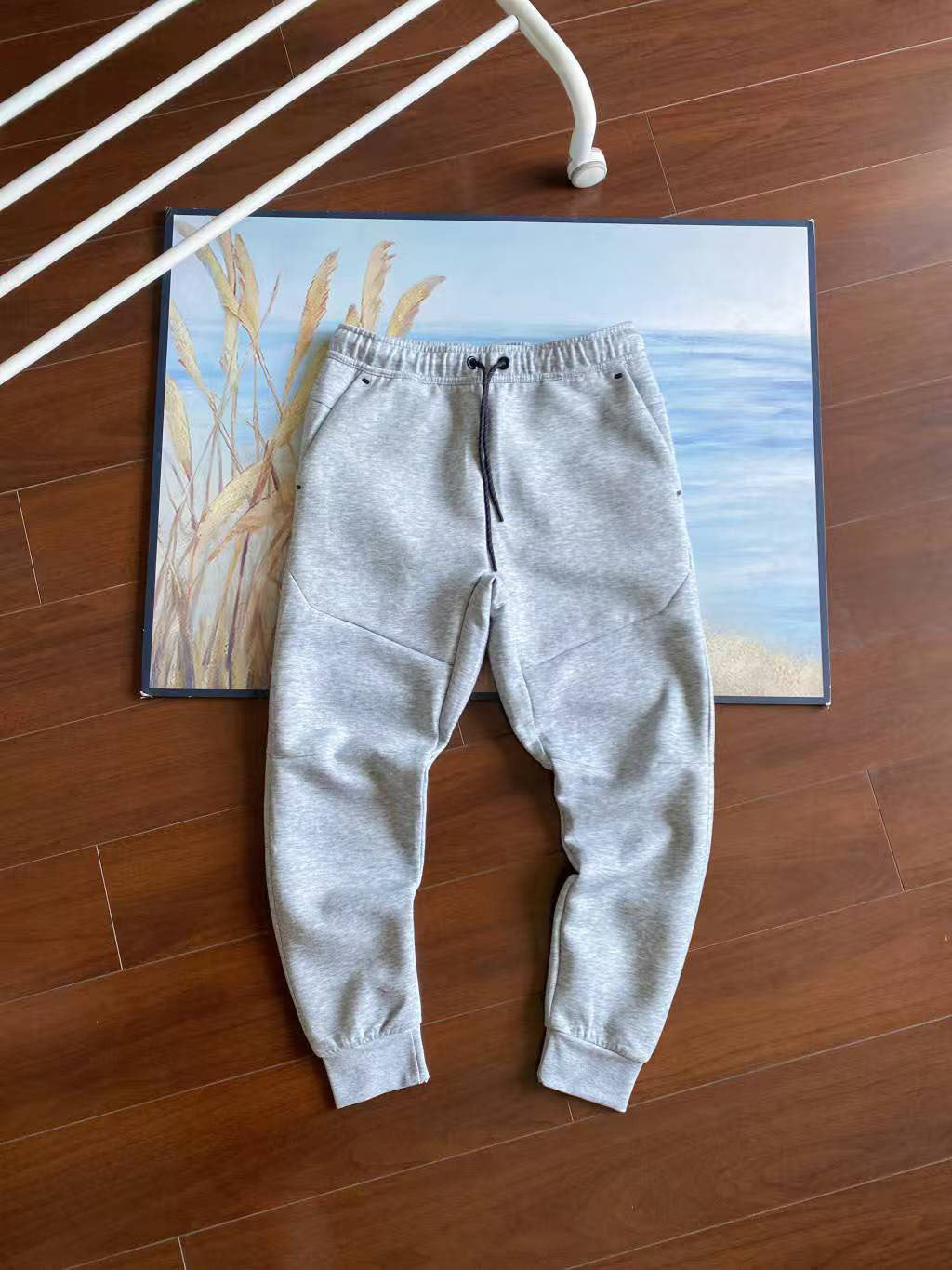 Light gray heather sweatpants laid flat on a wooden floor, styled over a framed beach painting with a white metal chair leg visible in the top left.