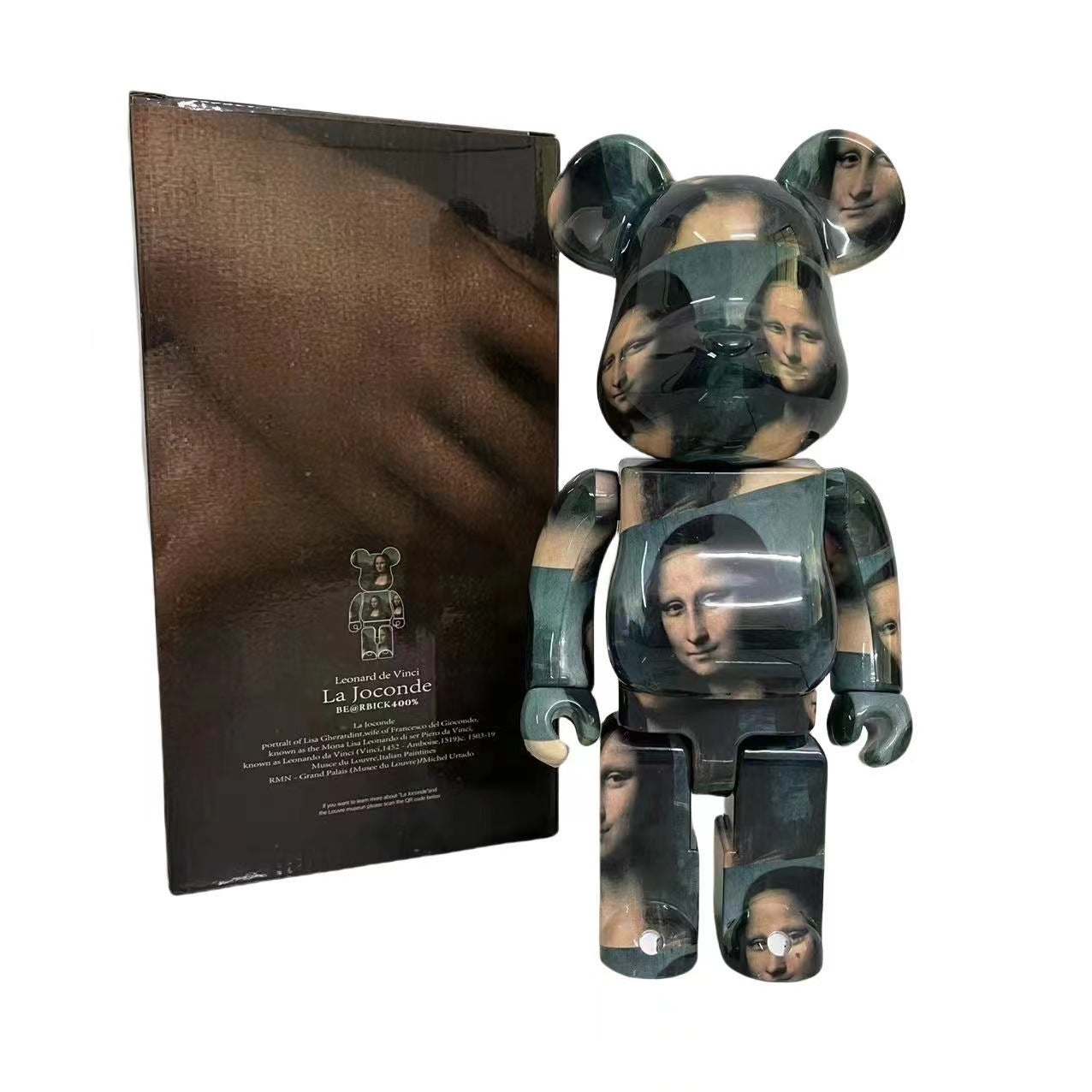 A collectible bear-shaped figure standing in front of its box, featuring a glossy finish with an all-over print of Leonardo da Vinci's Mona Lisa. The figure has dark teal-green eyes and arms, with lighter beige skin tones on the head, shoulders, and hands.