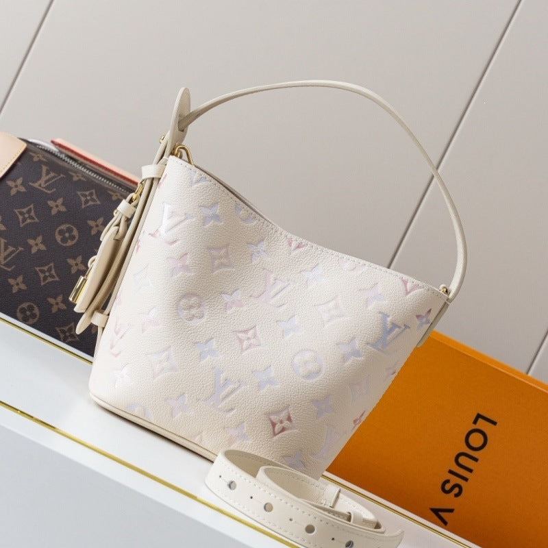 A Louis Vuitton monogram handbag in off-white leather with a top handle, displayed on a white shelf next to an orange Louis Vuitton box and another monogram bag.