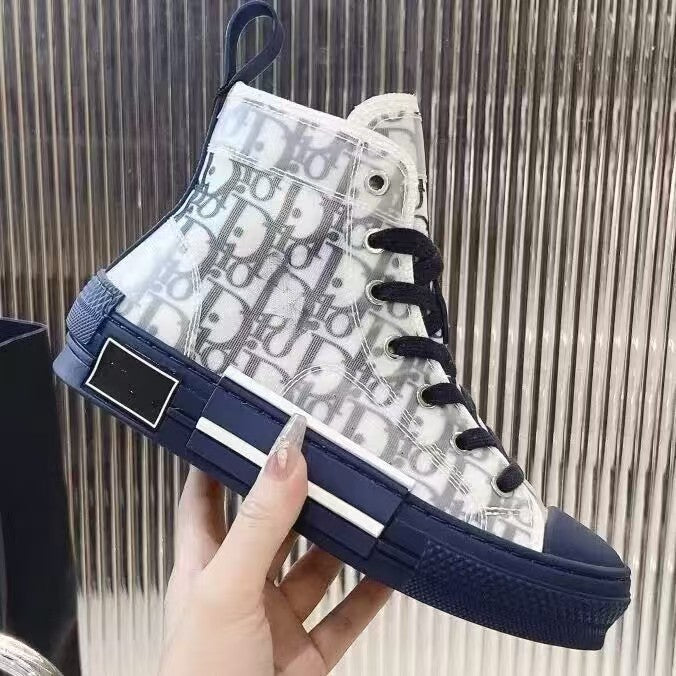 A hand holds a high-top sneaker featuring a white and navy blue Dior monogram canvas upper, navy laces, and a navy sole with a white stripe, set against a metallic ribbed background.