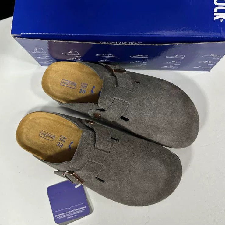 Pair of gray suede shoes with a brown sole on a white surface, with a blue box in the background.