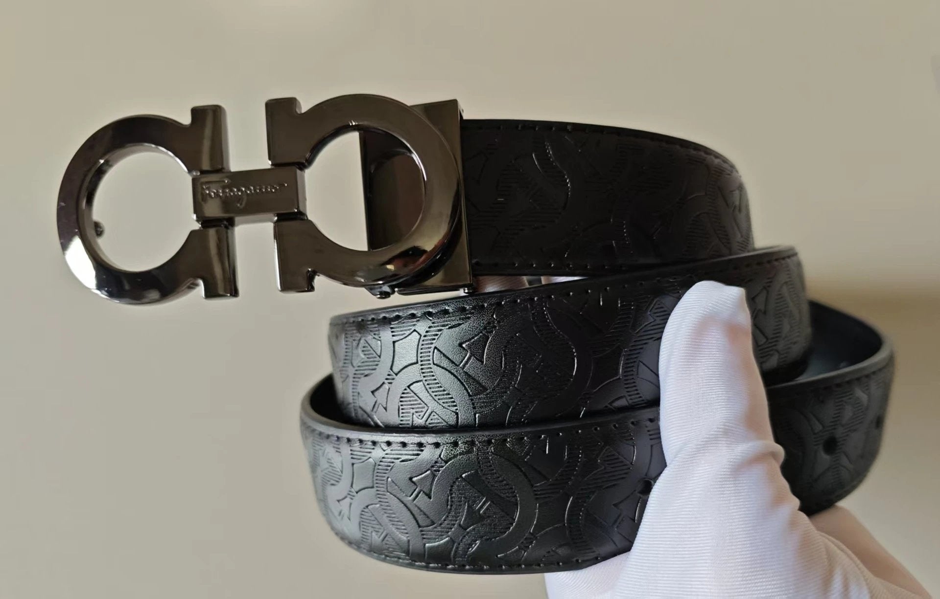 Black leather belt with a prominent logo held by a gloved hand against a neutral background