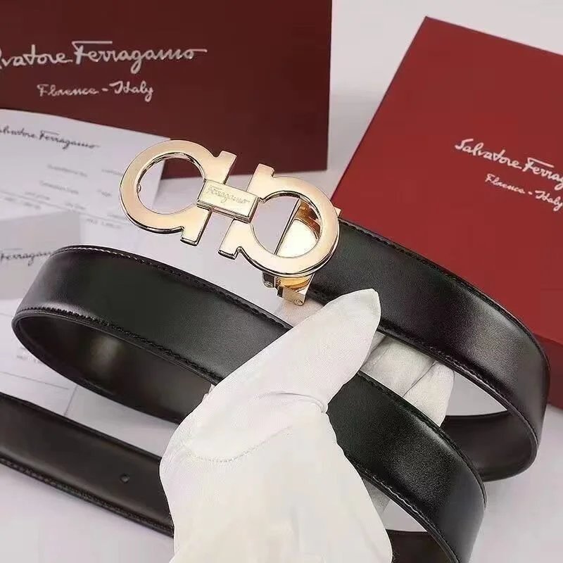Black Salvatore Ferragamo belt with gold buckle held by a gloved hand, with brand packaging in the background.