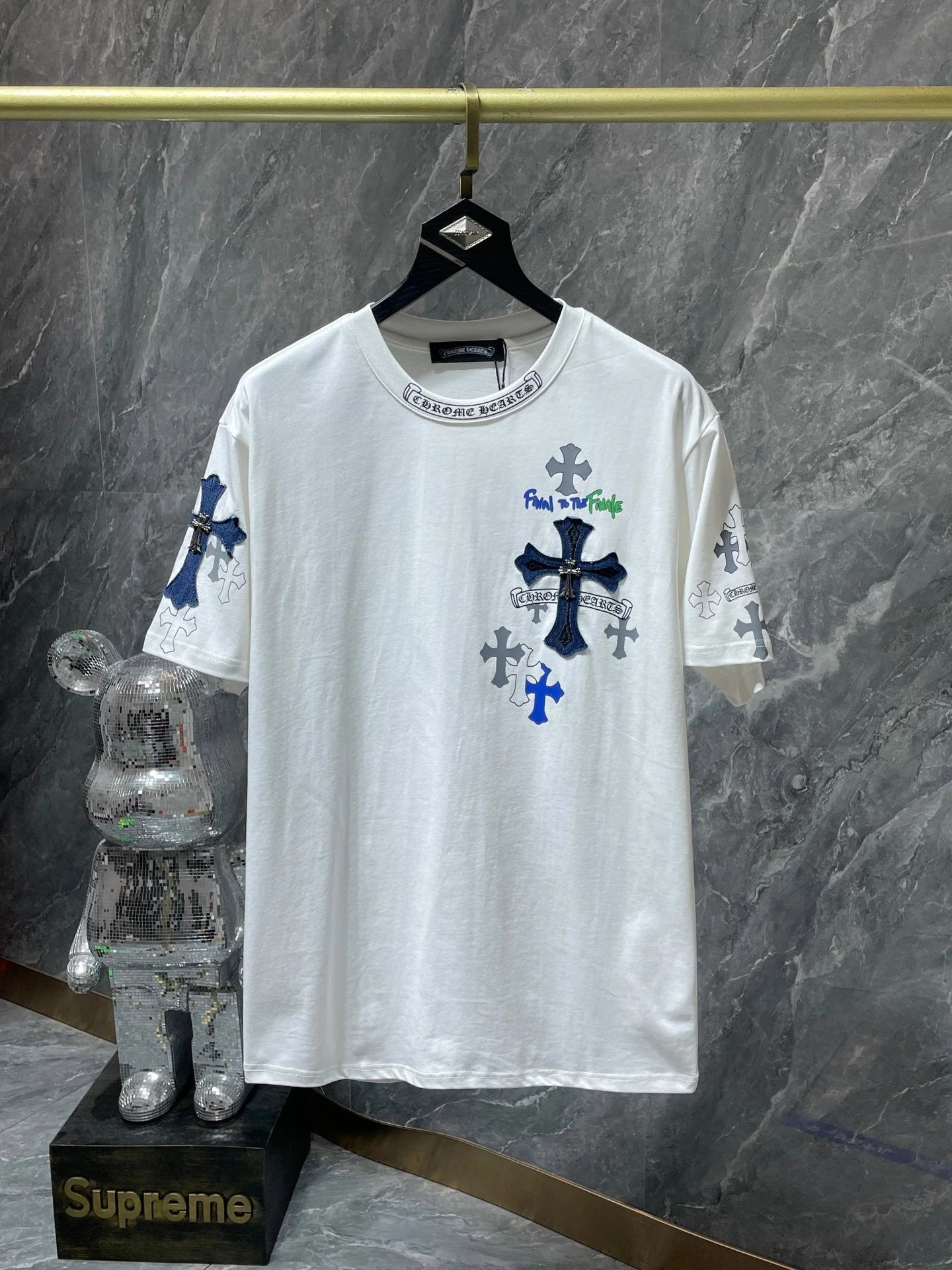 White t-shirt with cross designs on a hanger against a marble wall, with a Supreme branded rabbit figure below.