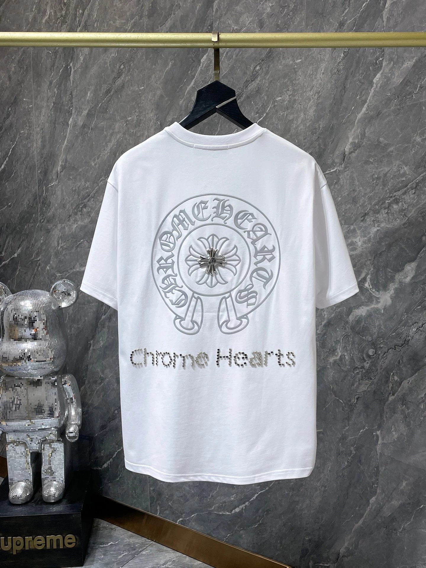 White t-shirt with 'Chrome Hearts' logo on a hanger against a marble wall.