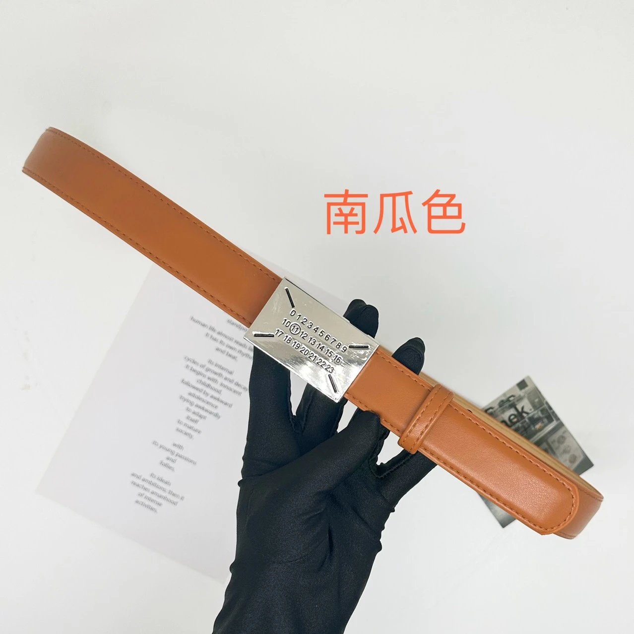 A tan leather belt with a silver-toned metal buckle held by a black gloved hand. The image features Chinese text '鱼菌吉' (pumpkin color) in orange, and a white paper insert is visible in the background.