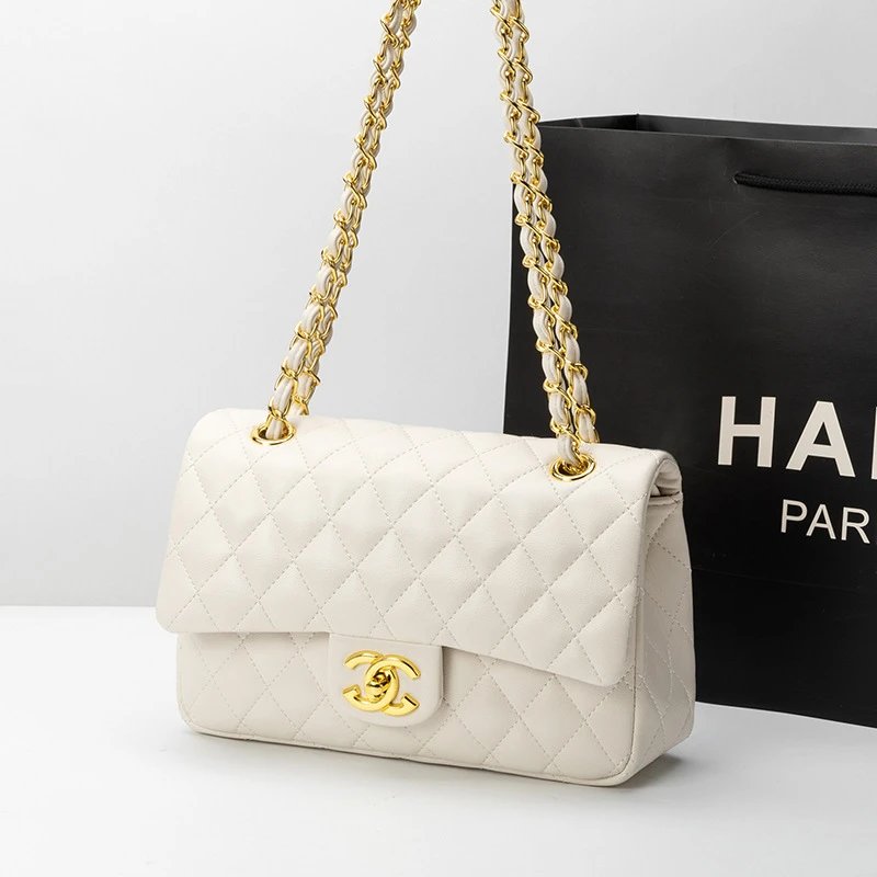 A white quilted Chanel flap bag with gold-tone hardware and chain straps, shown in front of a black Chanel shopping bag on a light gray background.