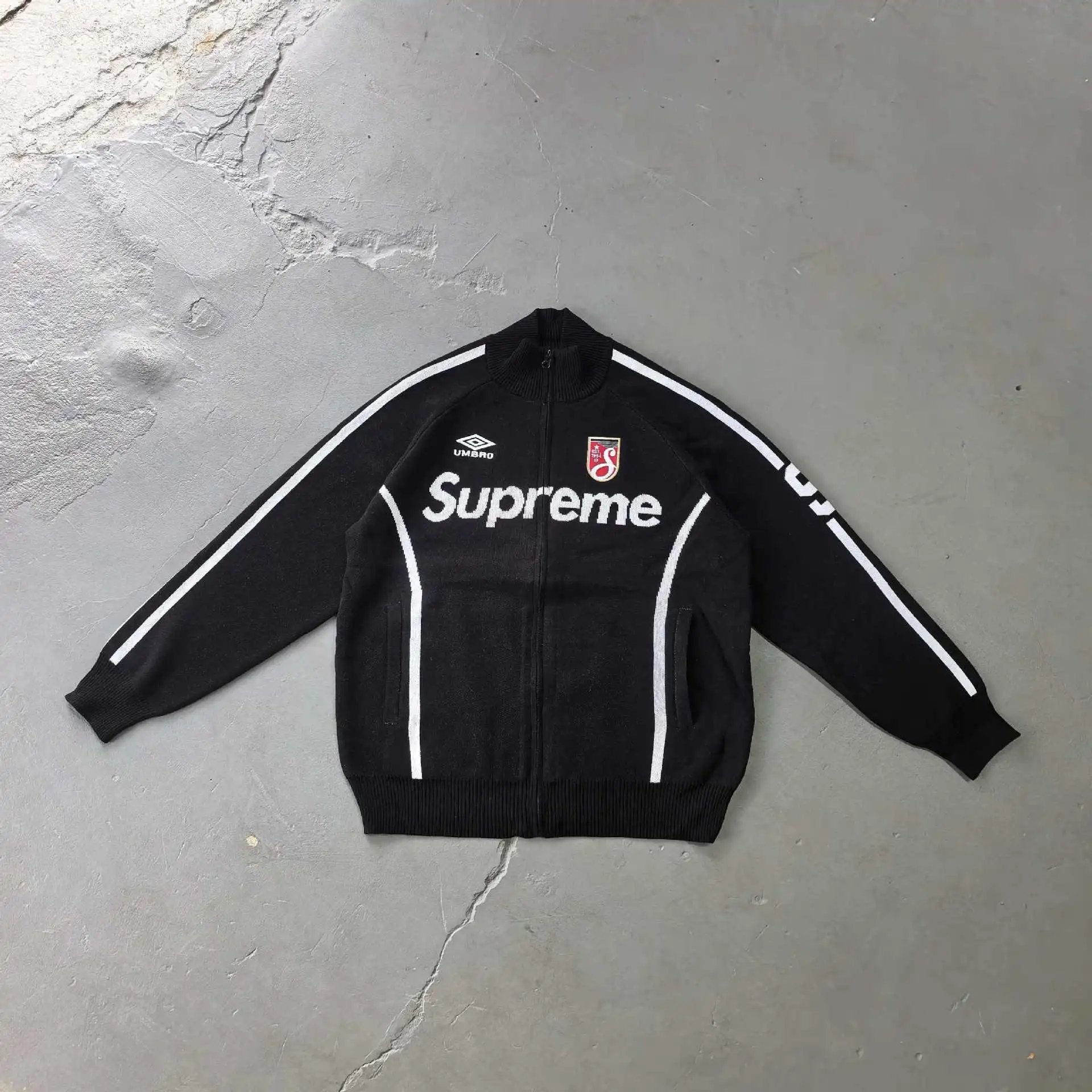 A black Supreme zip-up track jacket with white stripes and branding, laid flat on a textured gray concrete floor.
