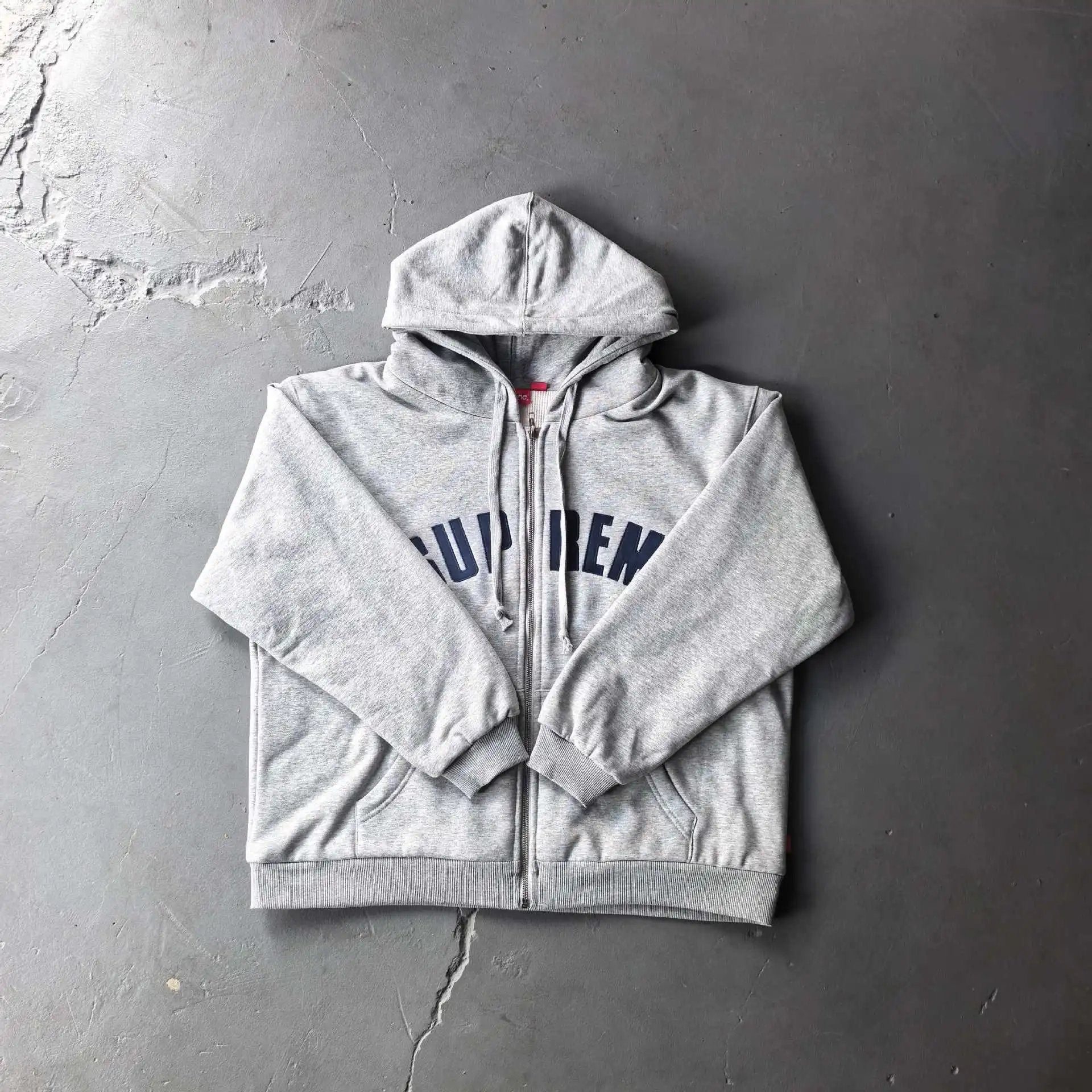 A light gray heather zip-up hoodie with 'SUPREM' printed in navy blue across the chest, laid flat on a textured gray concrete surface.