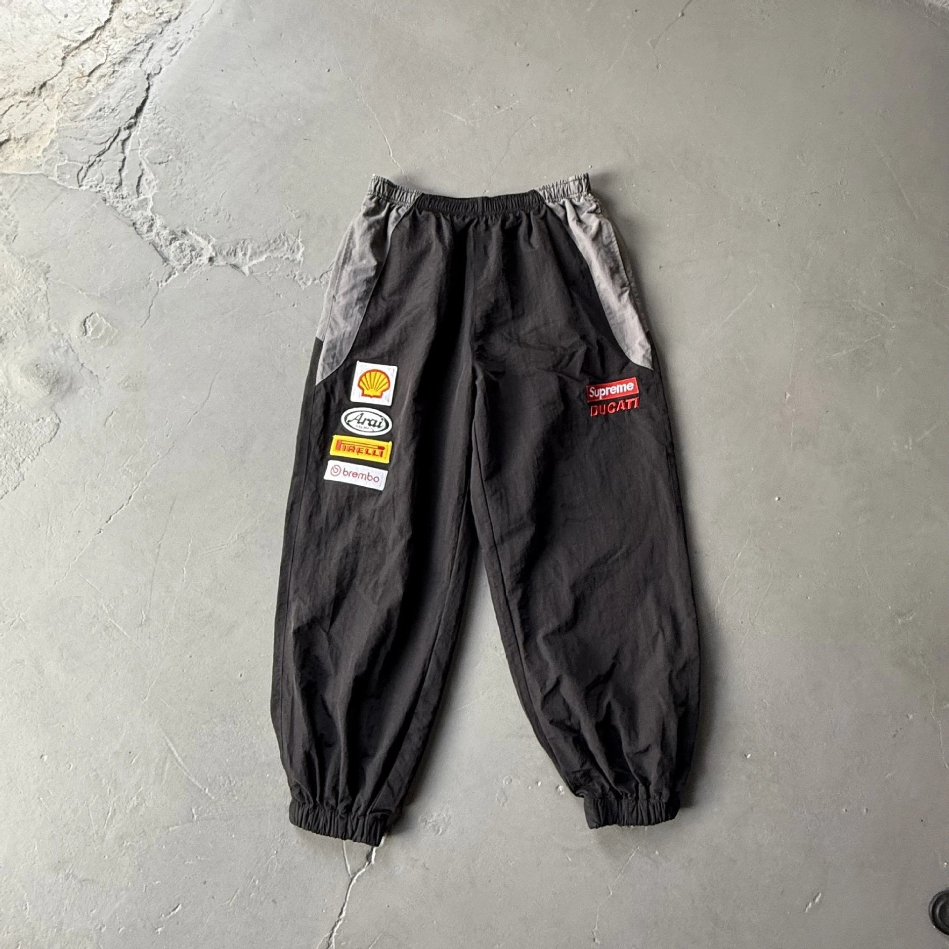 Black cargo jogger pants with gray side panels and multiple racing-style sponsor patches, laid flat on a gray concrete floor.