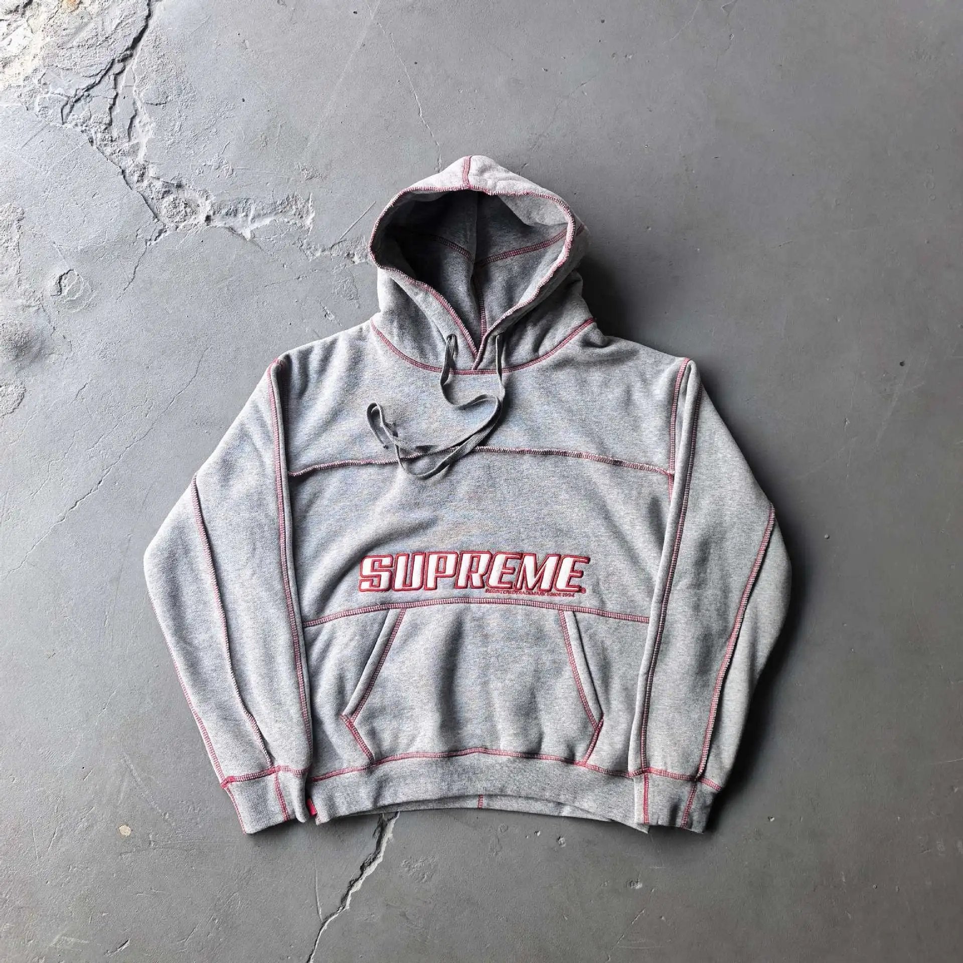 A heather gray Supreme hoodie with red contrast stitching and a red-and-white embroidered Supreme logo on the chest, laid flat on a textured gray concrete surface.