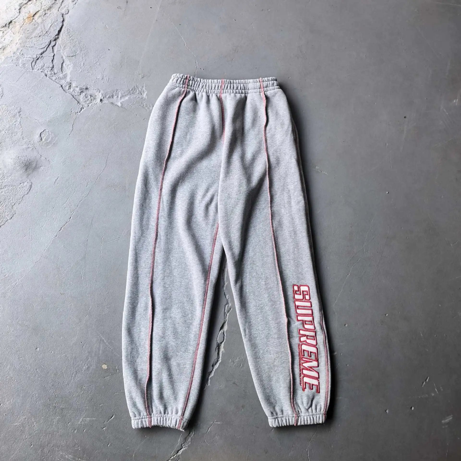 A pair of heather gray sweatpants with red contrast stitching and a red Supreme logo printed vertically on the left leg, laid flat on a gray concrete floor.