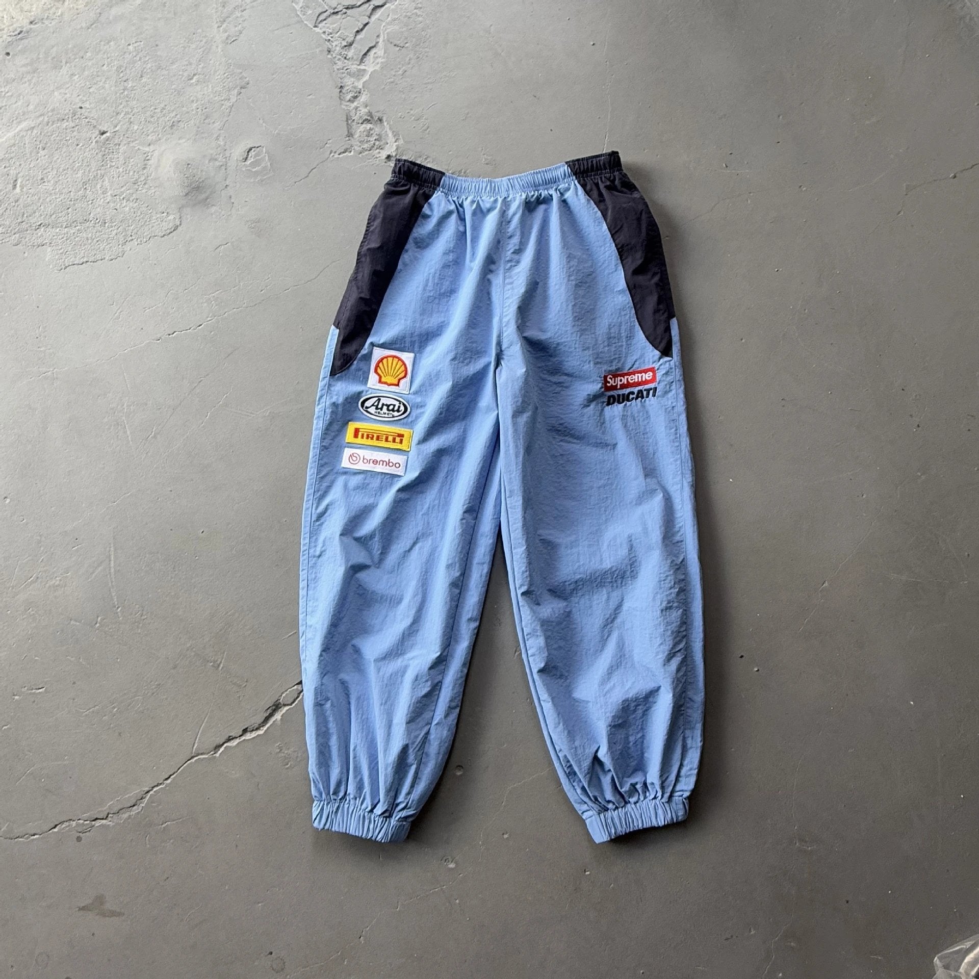 Light blue windbreaker track pants with black side panels, featuring sponsor patches including Shell, Pirelli, and Ducati on the left leg, laid flat on a gray concrete floor.