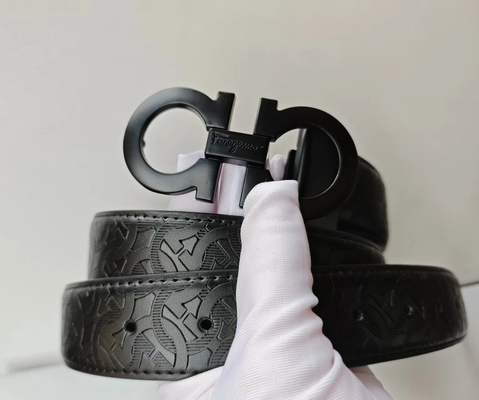Black leather belt with Ferragamo logo held by a gloved hand against a neutral background