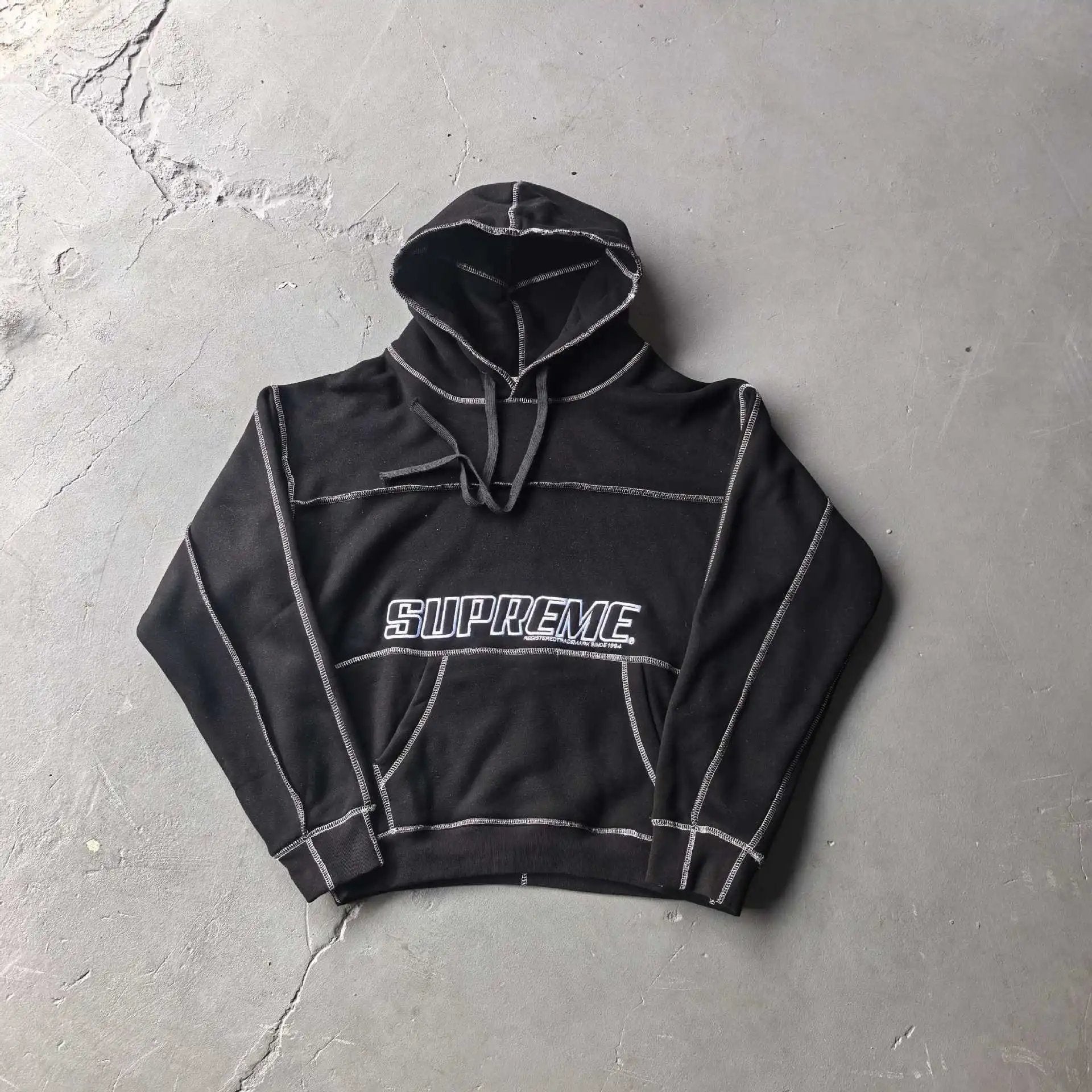 A black cropped hoodie with white contrast stitching and a white Supreme logo across the chest, laid flat on a textured gray concrete surface.