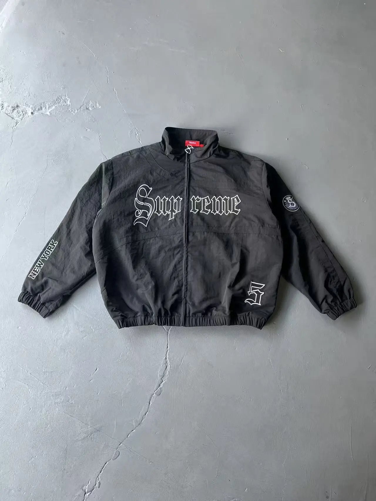 A black zip-up jacket with 'Supreme' written in gothic white lettering across the chest, laid flat on a gray concrete floor.