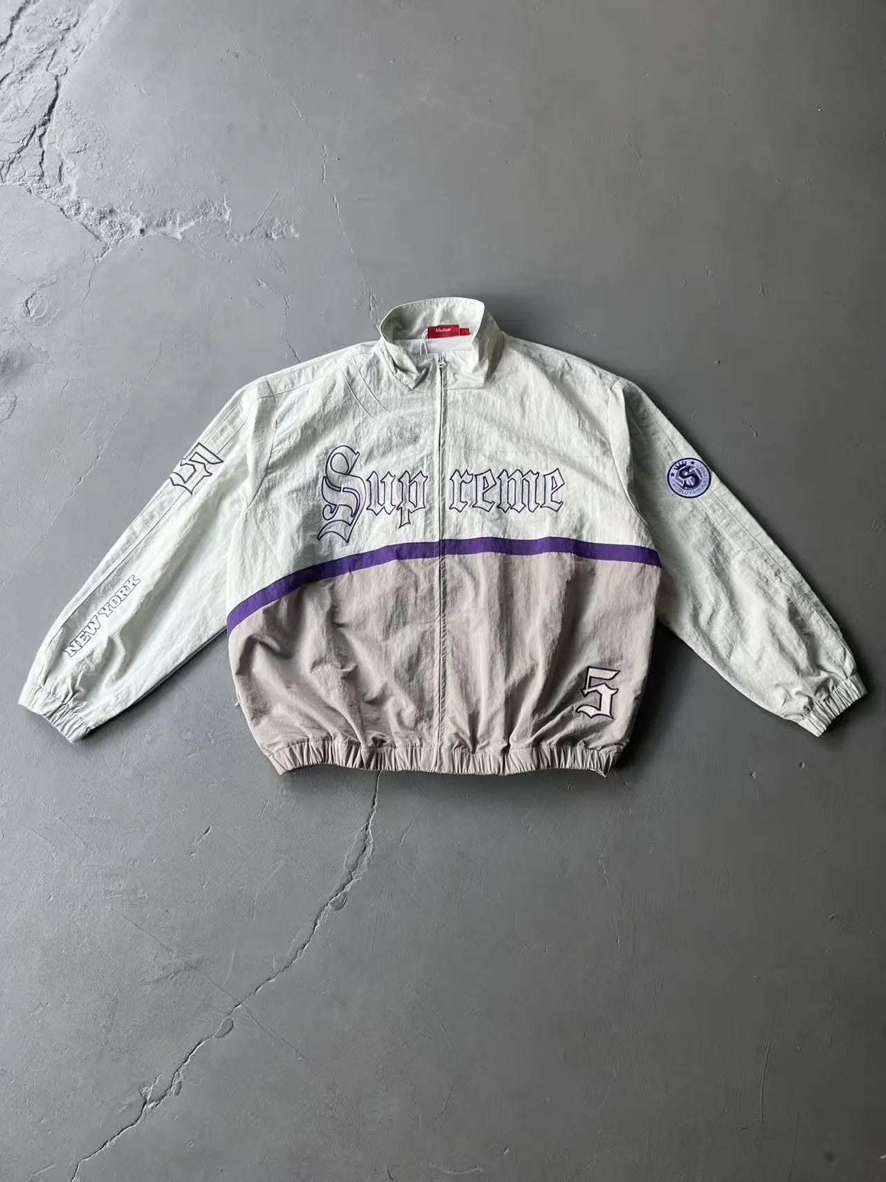 A Supreme windbreaker jacket laid flat on a gray concrete floor. The jacket features a cream upper body with purple gothic 'Supreme' lettering, a purple horizontal stripe, and a taupe lower body with a purple stripe.