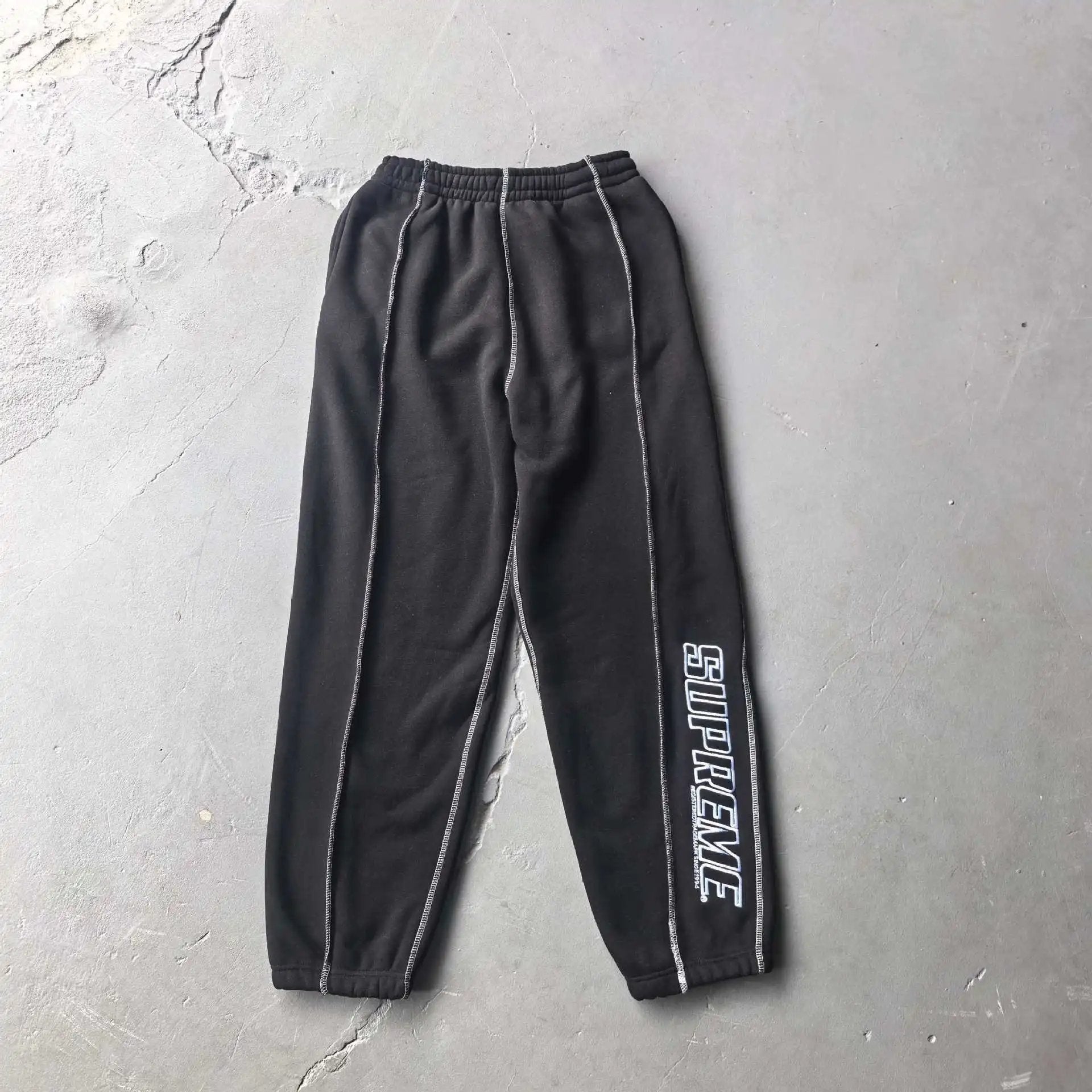 Black sweatpants with white contrast stitching and a white vertical 'STUDYPLAZZE' text graphic on the left leg, laid flat on a gray concrete floor.