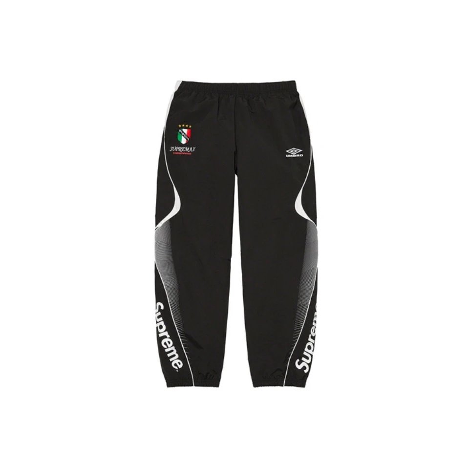 Black Supreme x Umbro track pants featuring white 'Supreme' text down the legs, a white Umbro logo on the right hip, and a green, white, and red Italian flag patch on the left hip.