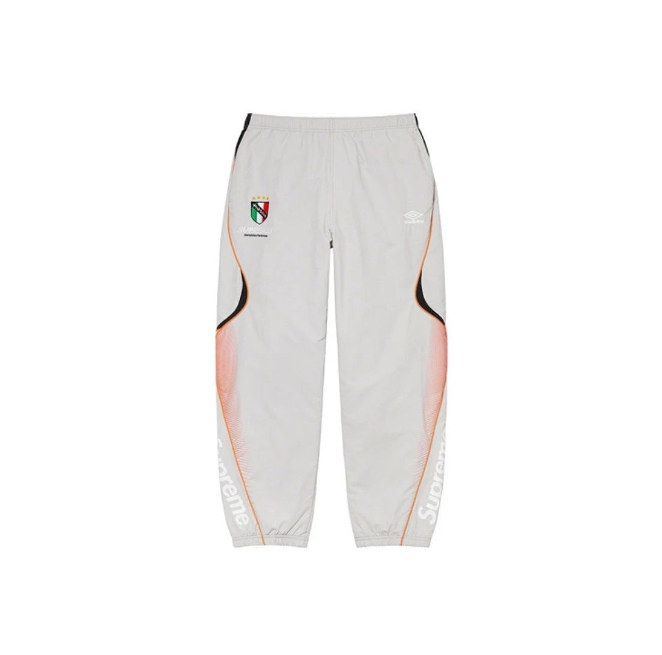 White Supreme x Inter Miami CF track pants featuring orange and black side accents, a team crest on the left chest, and 'Supreme' printed down the right leg.
