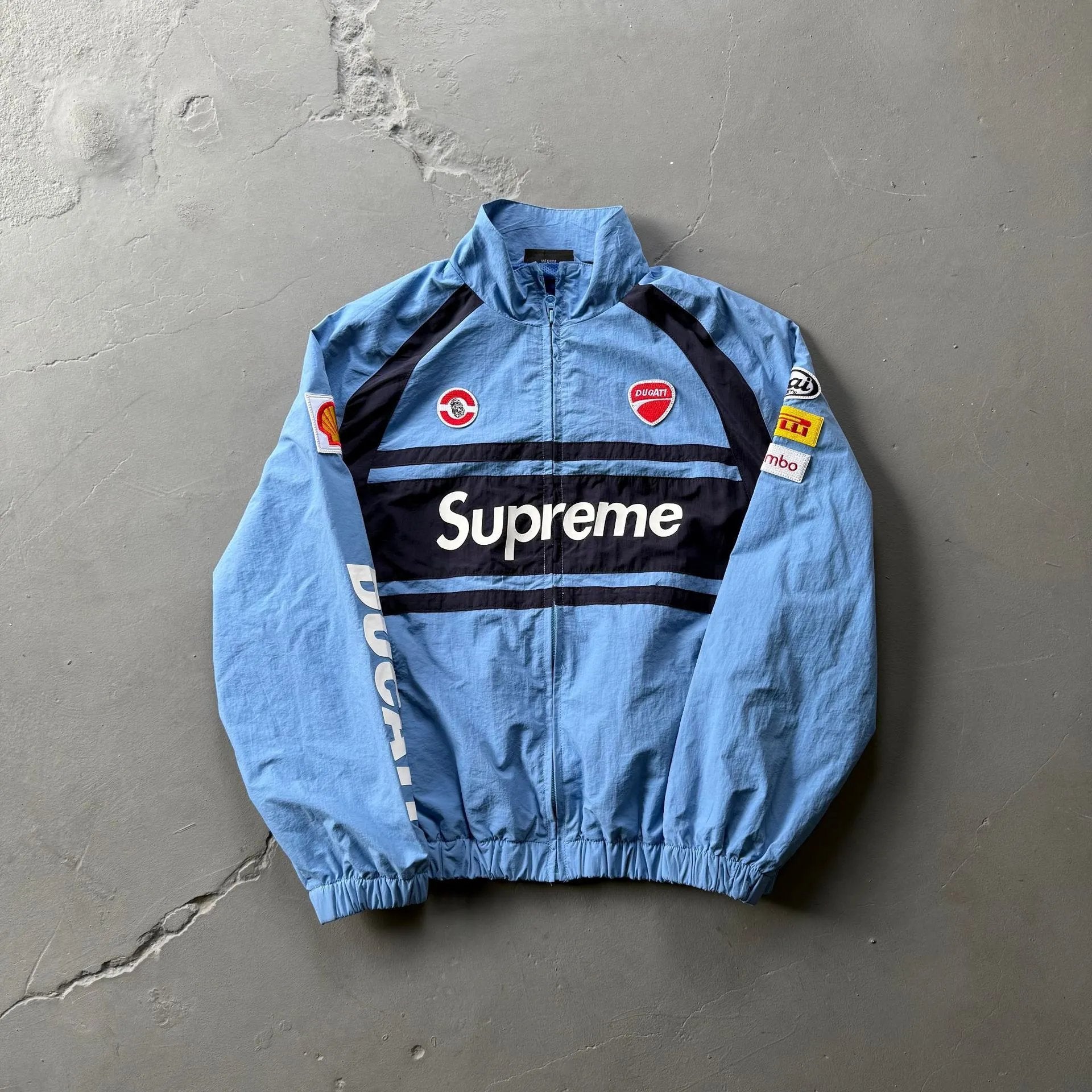 A light blue windbreaker jacket featuring a black chest stripe with 'Supreme' in white, Ducati branding on the sleeves, and various sponsor patches, laid flat on a gray concrete surface.