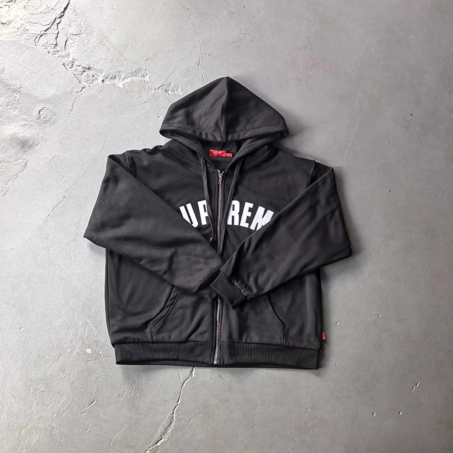 A black Supreme zip-up hoodie with white 'SUPREME' text across the chest, laid flat on a textured gray concrete surface.