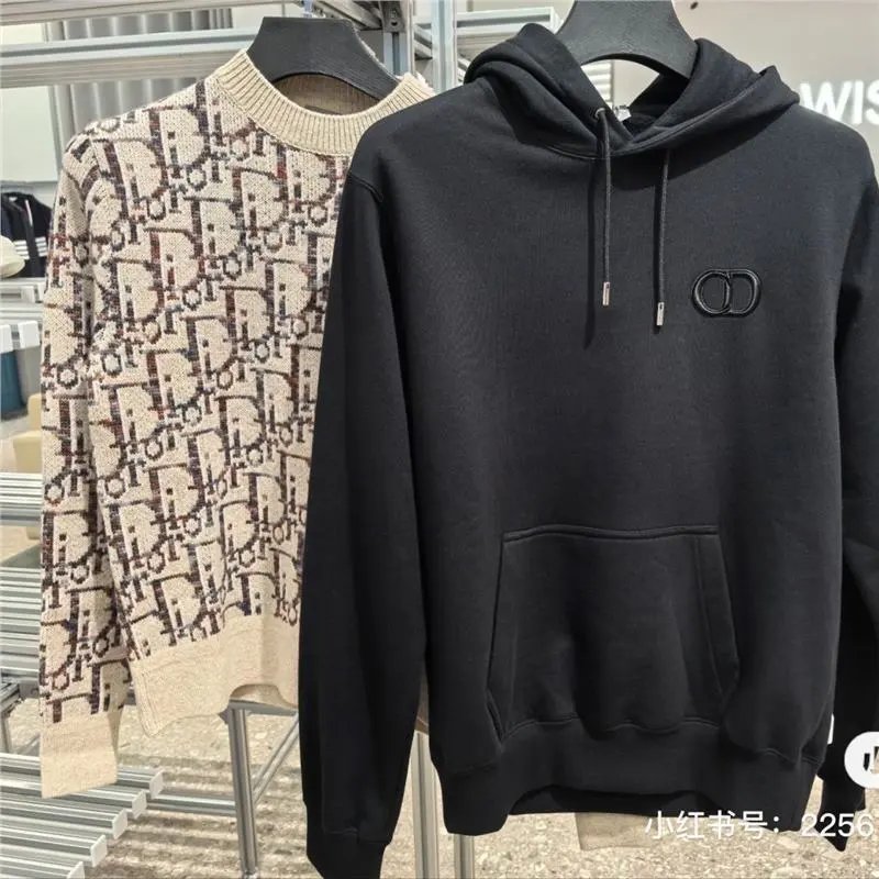 Two hooded knit sweaters hanging on black hangers in a retail setting. The foreground sweater is solid black with a small tonal double-ring logo on the chest and a front kangaroo pocket.