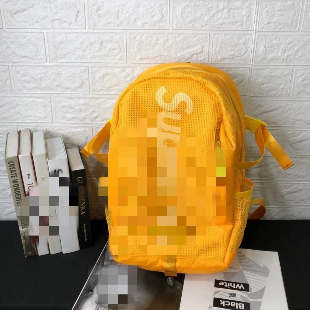 A bright yellow mesh backpack with a pixelated block overlay covering the front logo area, displayed on a black tabletop against a white brick wall backdrop.