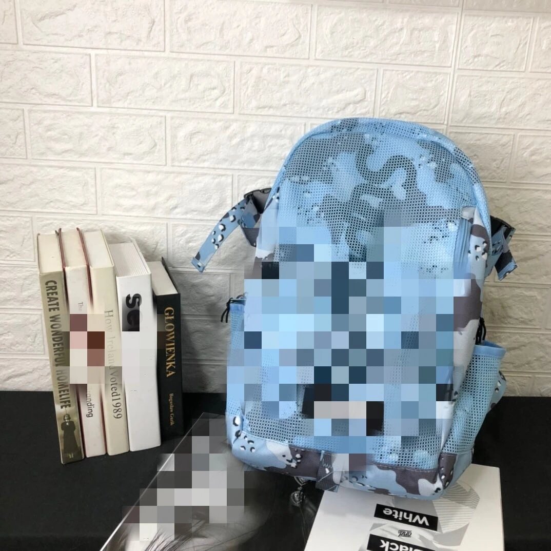A light blue and gray camouflage backpack with pixelated blurring over the front pocket, displayed on a black tabletop next to books against a white brick wall.