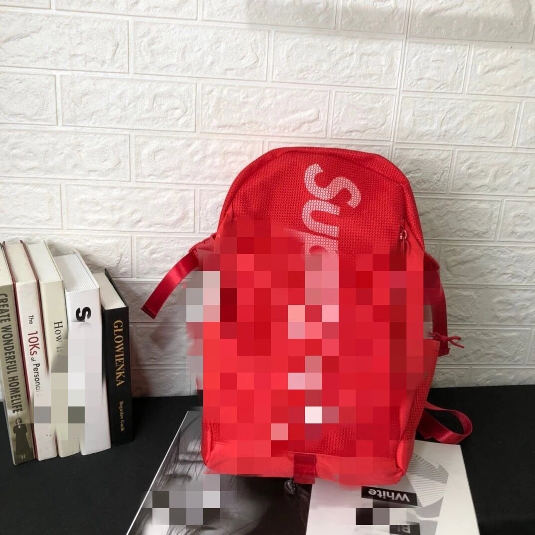 A red canvas backpack with a pixelated blur over the front logo area, displayed on a black tabletop against a white textured brick wall, next to a stack of books.