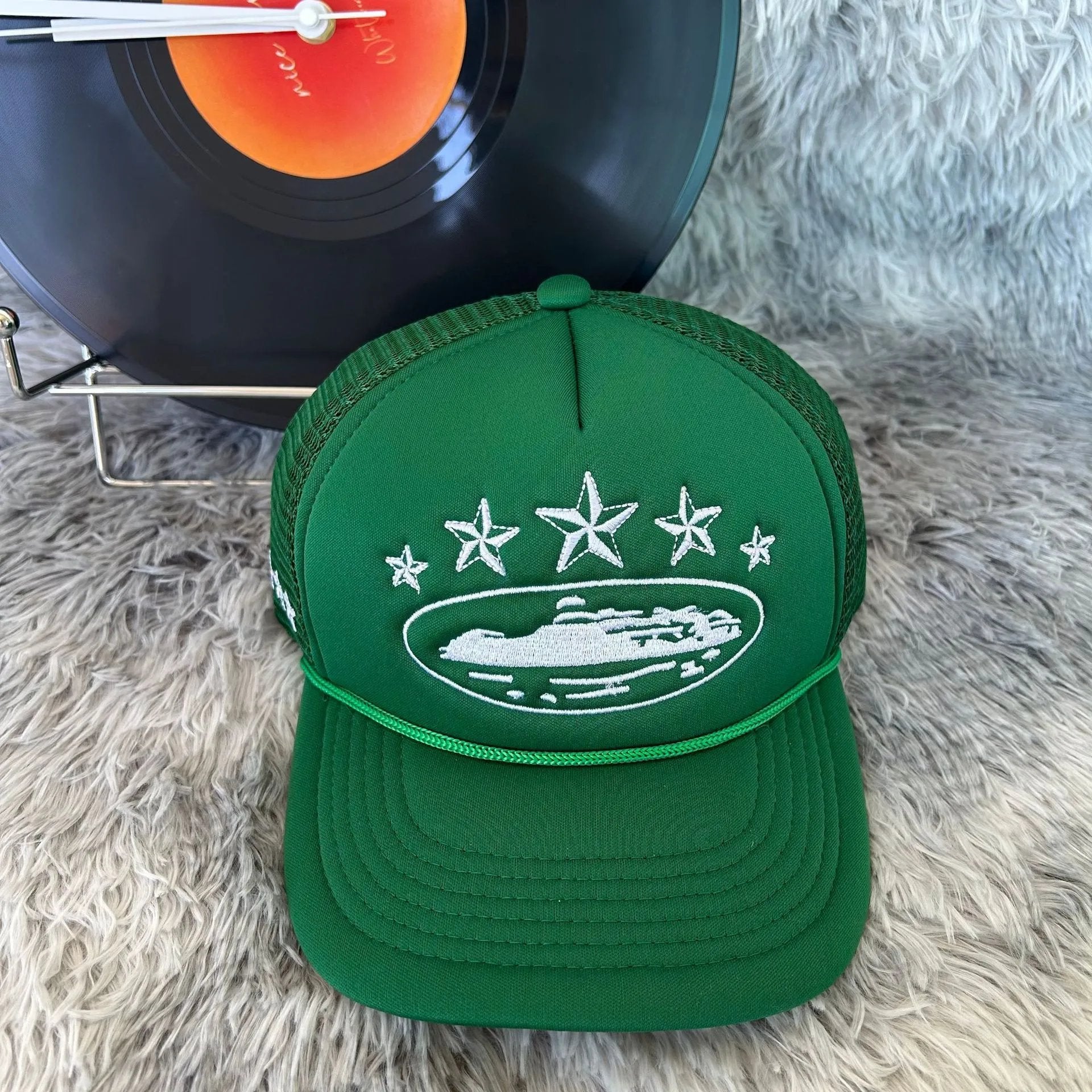 A green trucker-style cap with white embroidered stars and a ship graphic on the front, displayed on a shaggy rug with a vinyl record in the background.