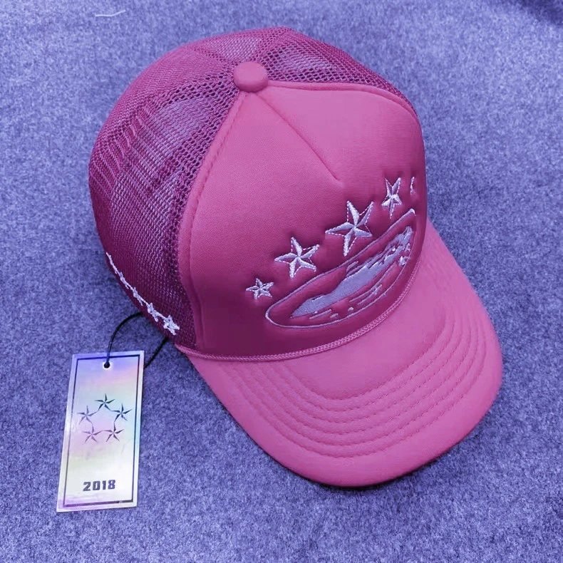A pink trucker-style baseball cap with mesh back panels and white embroidered stars and a curved logo on the front, shown with a hangtag on a textured blue surface.