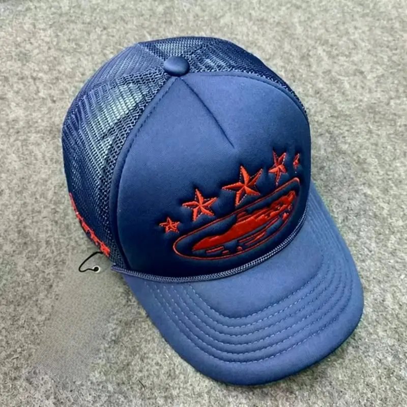 A blue trucker-style baseball cap with orange embroidered stars and a red car logo on the front, photographed on a gray textured surface.
