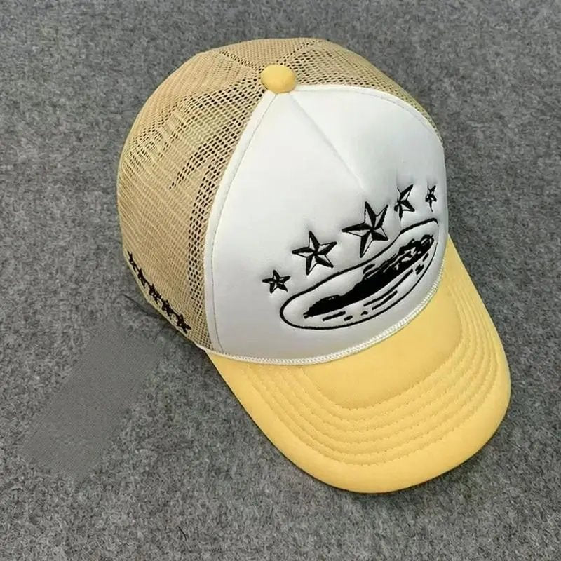 A yellow and beige trucker hat with a white front panel, featuring a black graphic of a hot dog with stars above it, photographed on a gray felt surface.