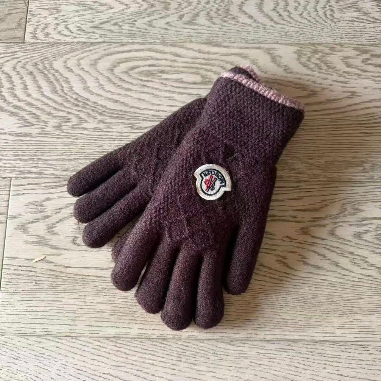 A pair of dark plum purple knit work gloves with a circular white logo patch on the back of one glove, displayed on a light wood grain surface.