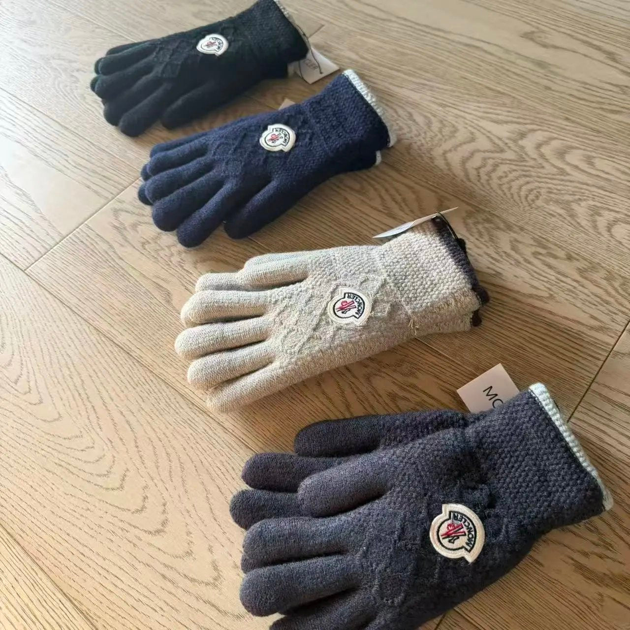 Four cable-knit winter gloves arranged diagonally on a light wood floor. The gloves come in four colorways: black, navy blue, cream, and dark gray.