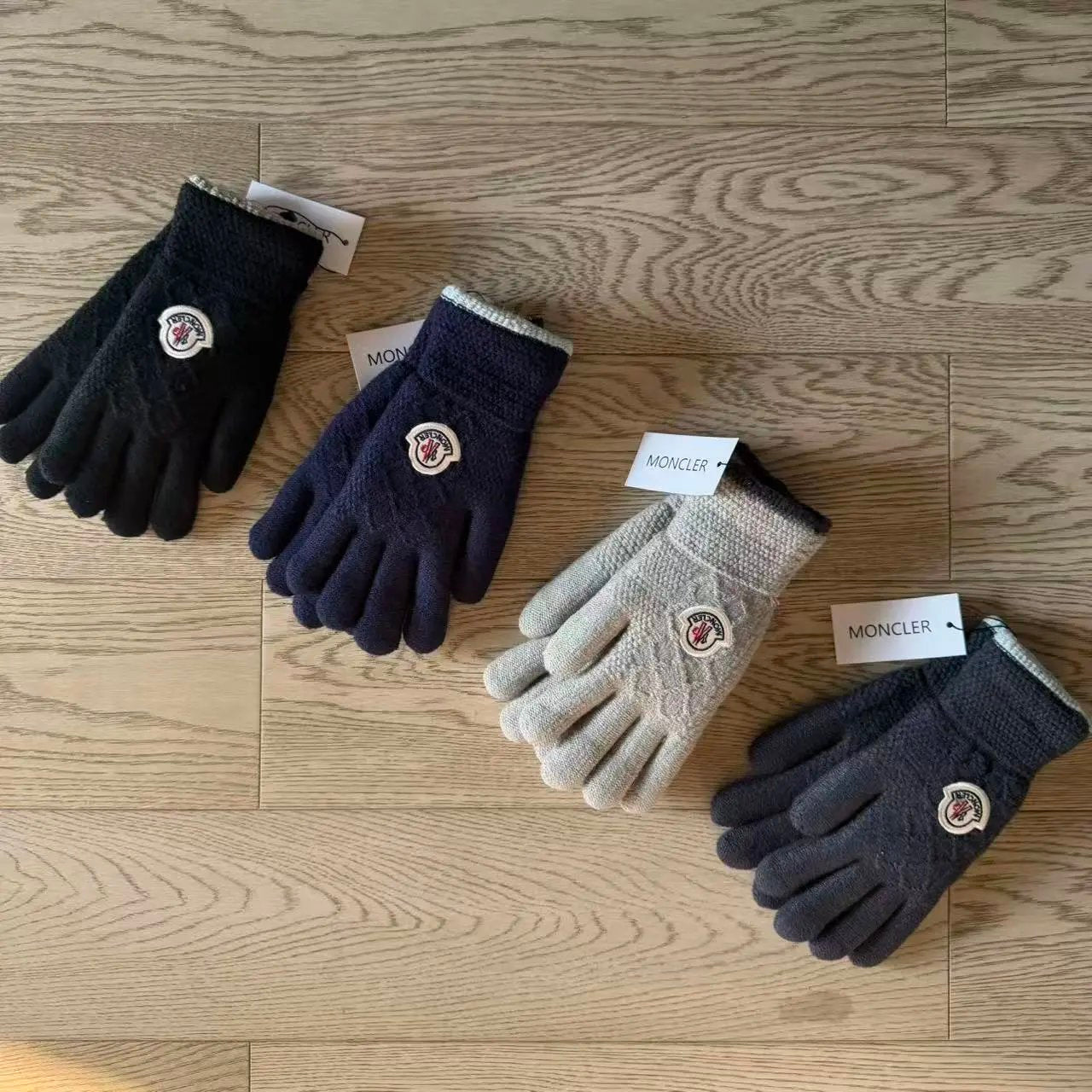 Four Moncler knit gloves in different colors (black, navy, light beige, and charcoal) arranged on a light wood floor, each with its brand tag visible.