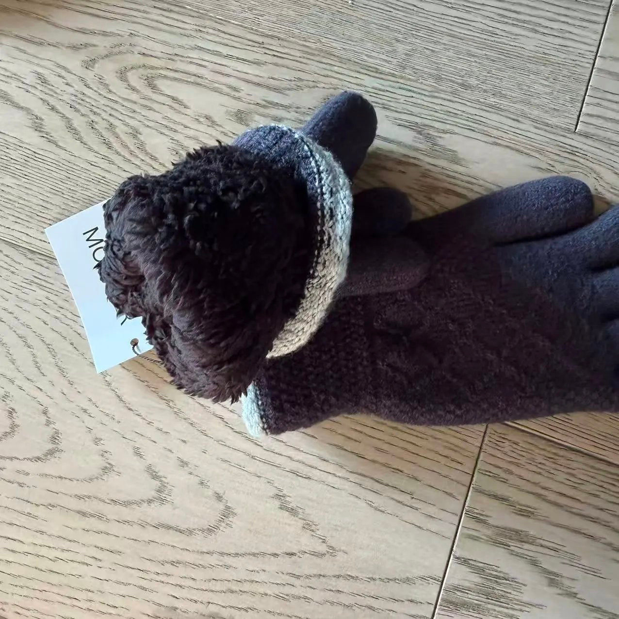 A pair of knit winter gloves in dark brown and black, with a striped cuff in brown, black, and white, resting on a light wood floor. A small paper tag is attached to one glove.
