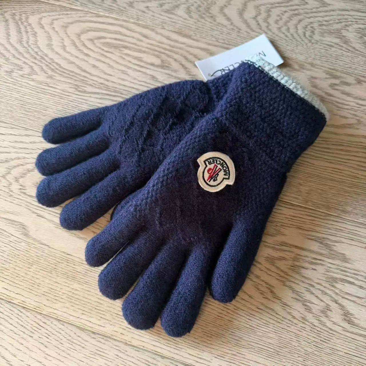 A pair of dark navy blue knit winter gloves with a circular embroidered Moncler logo patch on the back of one glove, resting on a light wood grain surface.