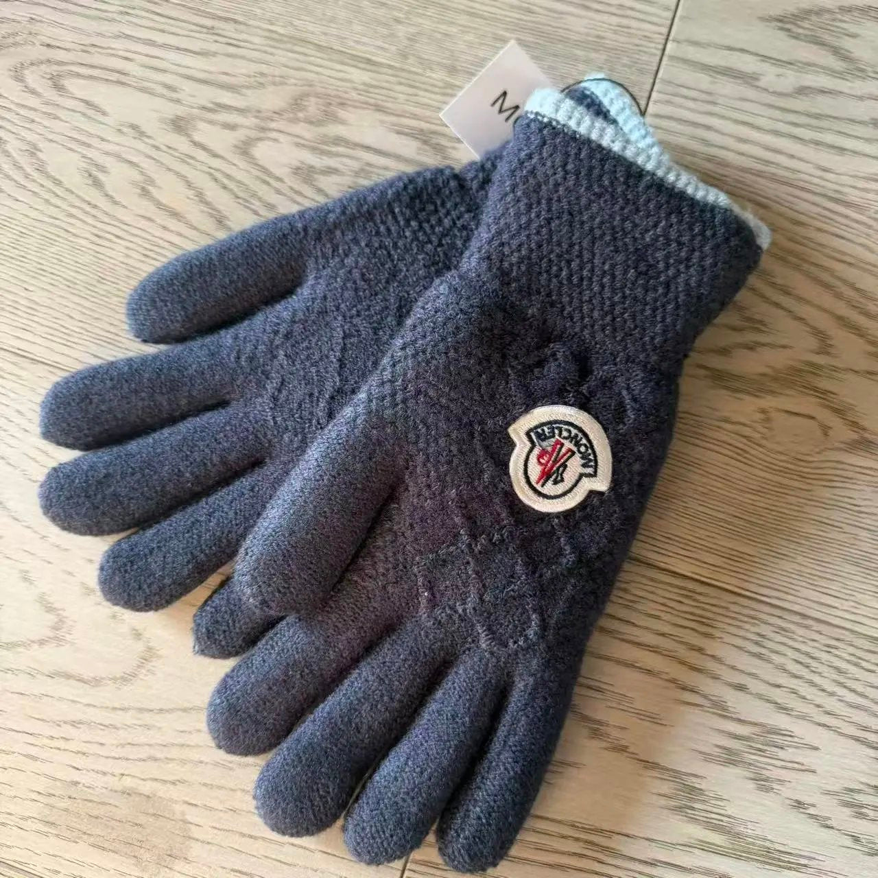 A pair of dark gray knit winter gloves with a light blue ribbed cuff, featuring a circular Moncler badge on the back of one glove, placed on a light wood grain surface.